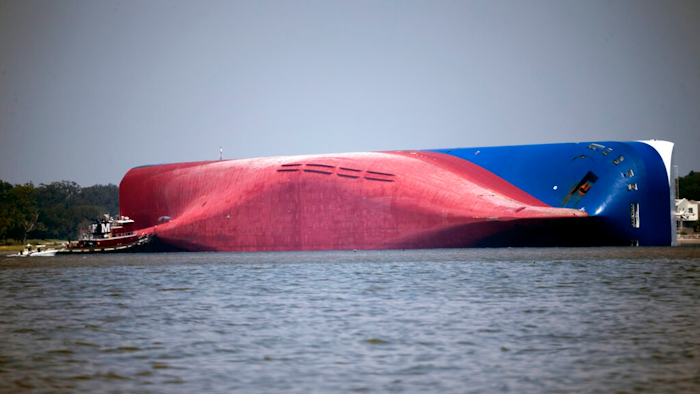 In this Sept. 9, 2019, file photo, a Moran tugboat nears the stern of the capsizing vessel Golden Ray near St. Simons Sound off the coast of Georgia. The salvage team is seeking a federal permit to surround the shipwreck with a giant mesh barrier to contain any debris when they cut the ship apart. Salvage workers coming to the Georgia coast to cut apart and remove the cargo ship that overturned will be isolated at a nearby resort to protect them from the coronavirus, officials said Tuesday, Sept. 8, 2020.