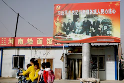 In this Sept. 20, 2018, file photo, an Uyghur woman uses an electric-powered scooter to fetch school children as they ride past a picture showing China's President Xi Jinping joining hands with a group of Uighur elders.