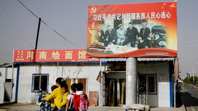 In this Sept. 20, 2018, file photo, an Uyghur woman uses an electric-powered scooter to fetch school children as they ride past a picture showing China's President Xi Jinping joining hands with a group of Uighur elders.
