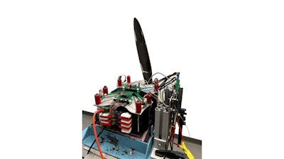 A battery sits on a wooden structure, hooked up to a propeller via exposed wires and electronic boards.