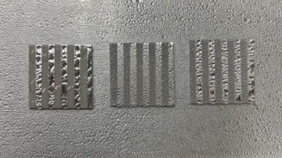 Aluminum coupon samples patterned with alternating stripes of smooth (lighter) and etched (darker) topographies display contrasting wettability.