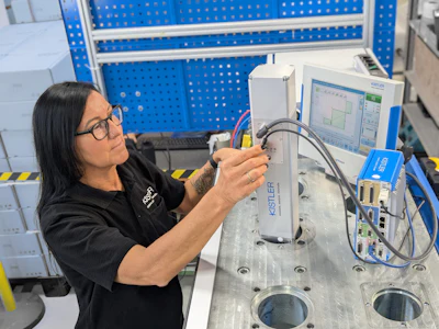 Female technician in glasses assembling or testing electronic equipment at industrial workstation with blue pegboard background
