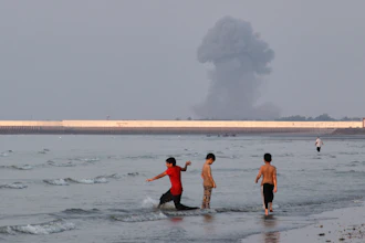 Three boys play in the shallow waters of the Strait of Hormuz as a plume of smoke rises from an explosion in the background off Bandar Abbas, Iran, July 13, 2026.
