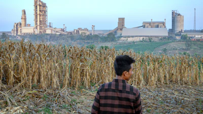 A farmer stands on a corm field as a cement factory seen in the background in Kendeng Mountains in Rembang, Central Java, Indonesia, Friday, June 19, 2026.