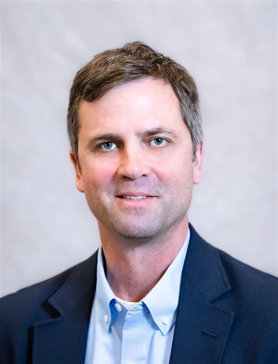 Professional headshot of a man in dark blazer and light blue shirt against neutral background