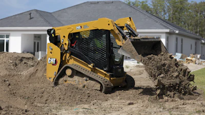 Yellow CAT 259 compact track loader moving soil at residential construction site with white house in background