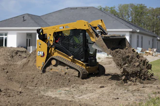 Yellow CAT 259 compact track loader moving soil at residential construction site with white house in background