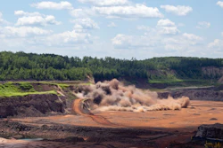 Heavy excavator creating dust cloud while mining in quarry with forested hillside backdrop