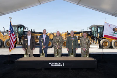 Six officials stand at podium with Northrop Grumman logo, flanked by American and California flags, with heavy equipment in background