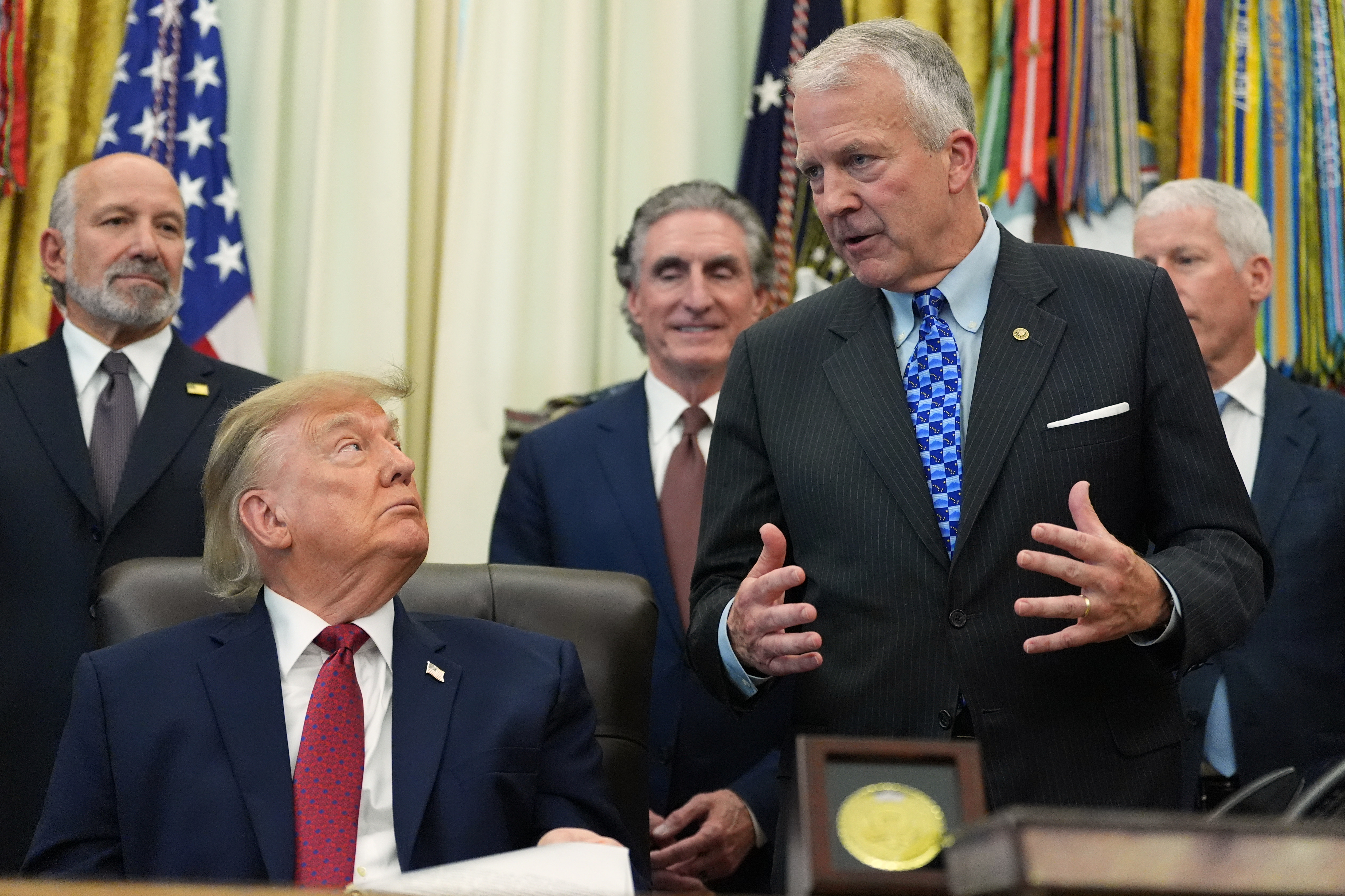 Formal meeting in the Oval Office with government officials and American flags in background