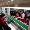 Electronics factory workers in maroon uniforms assembling components at industrial workstations in a bright manufacturing facility.