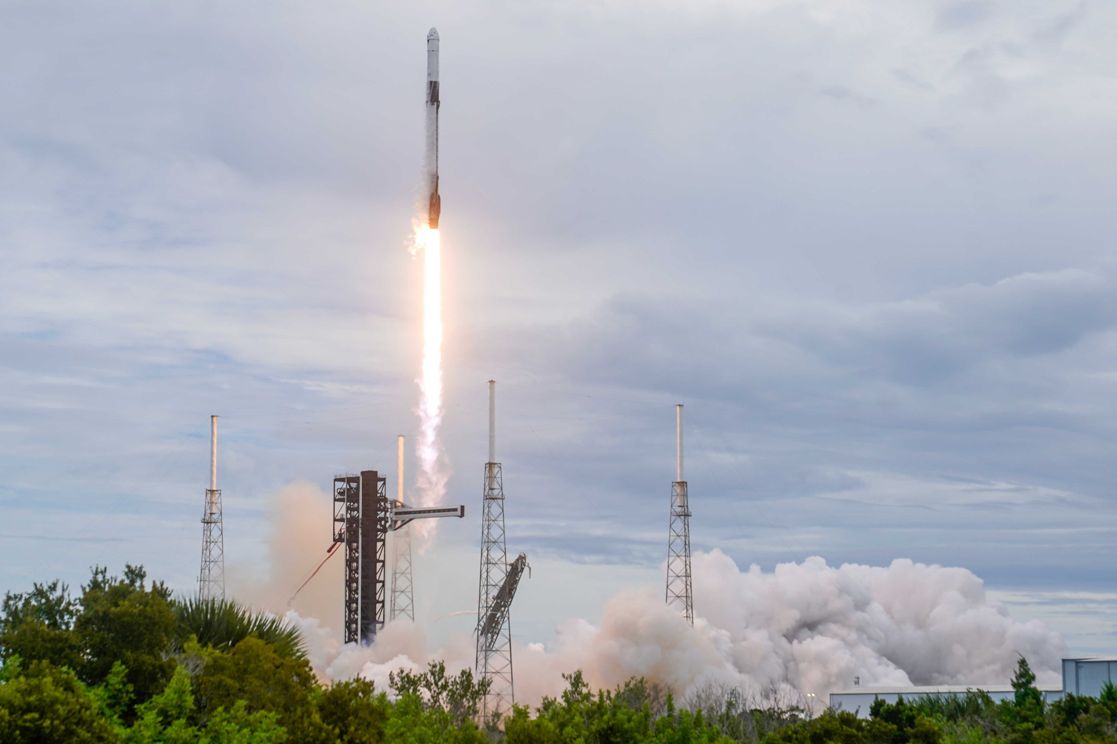 Rocket launching vertically with bright engine flames against cloudy sky and launch pad infrastructure visible below