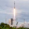 SpaceX Falcon 9 rocket launches from coastal launch pad with bright engine flames and white smoke clouds against cloudy sky