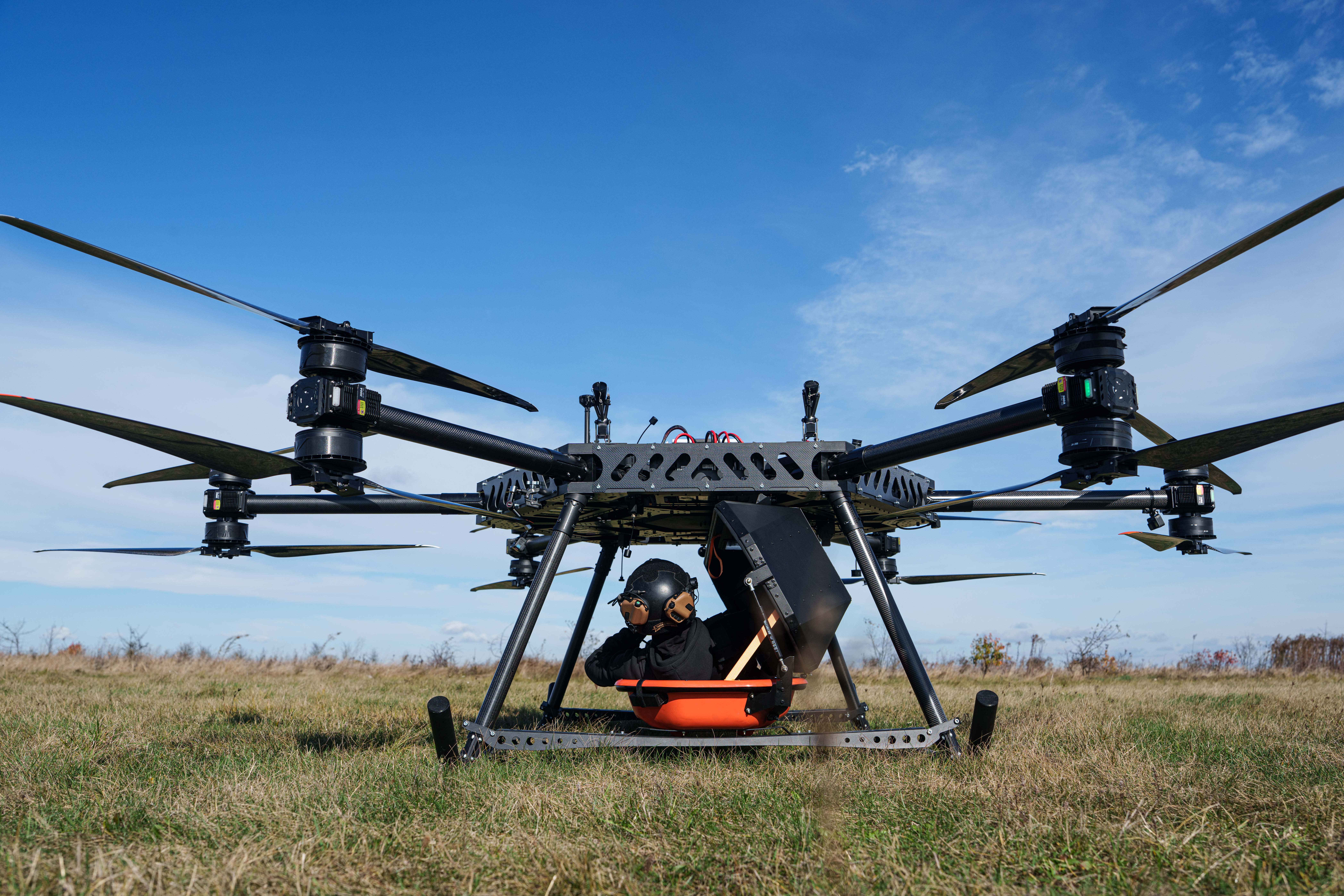 Large agricultural drone with orange payload container hovering above dry field under clear blue sky