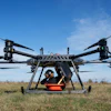 Large agricultural drone with orange payload container hovering above dry field under clear blue sky