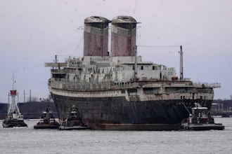 Large cargo ship assisted by tugboats in port waters with industrial twin smokestacks visible