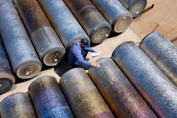 Worker in blue surveying large industrial metal pipes arranged on sandy ground from above