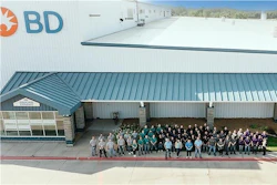 Large group of BD company employees standing together outside a modern industrial facility with white and teal metal roofing.