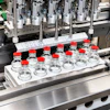 Pharmaceutical filling machine dispensing liquid into clear glass vials with red caps on production line