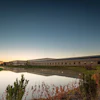 Modern industrial building complex reflected in calm water at dusk with autumn vegetation in foreground