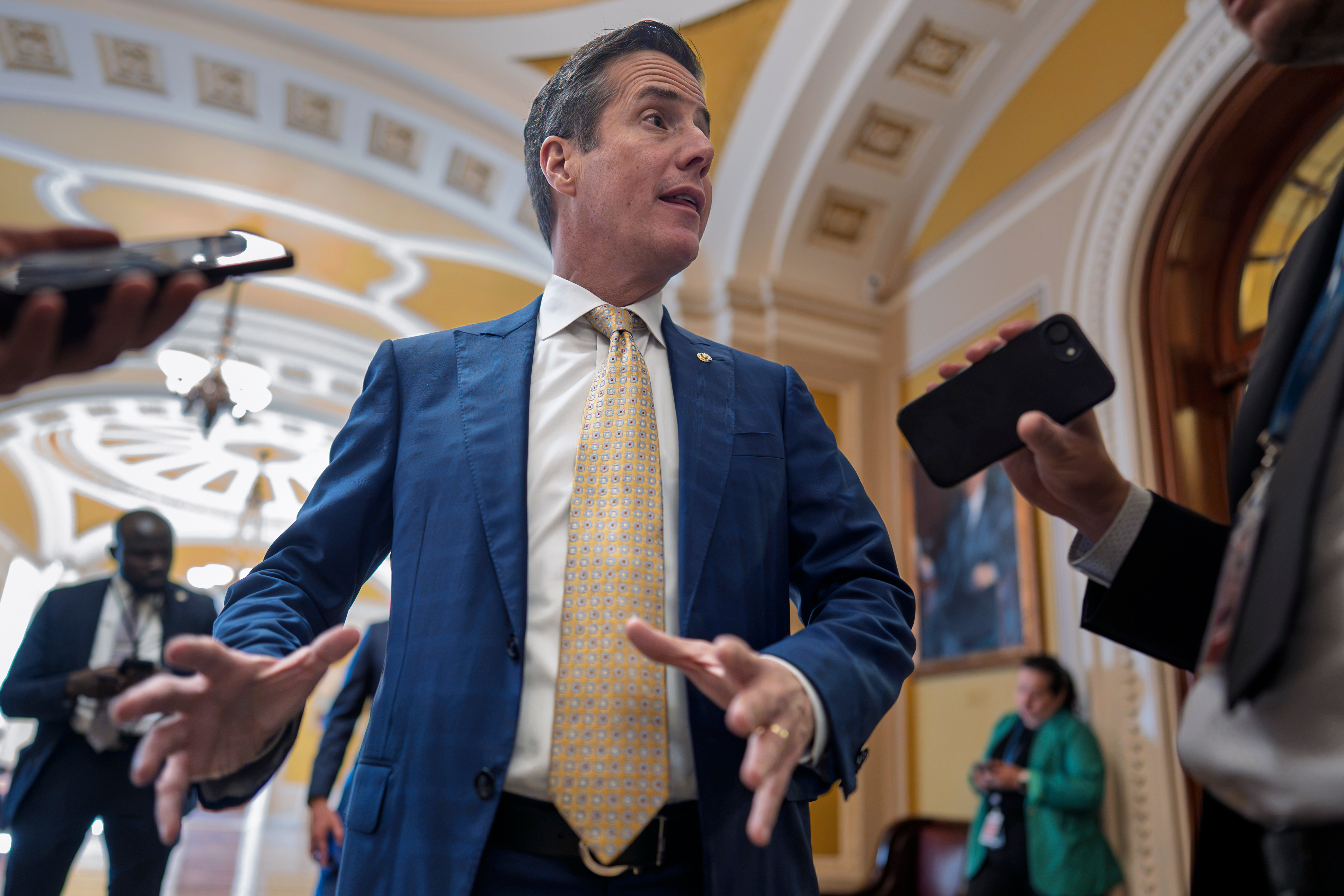 Man in blue suit and gold tie speaking to media in ornate government building corridor