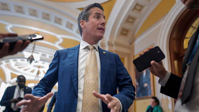 Man in blue suit and gold tie speaking to media in ornate government building corridor