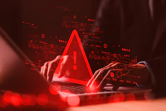 Red warning alert symbol glowing above laptop with hands, surrounded by digital data icons and circuit patterns on dark background