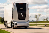White autonomous semi-truck with black cabin parked on a paved road with green fields and cloudy sky in background