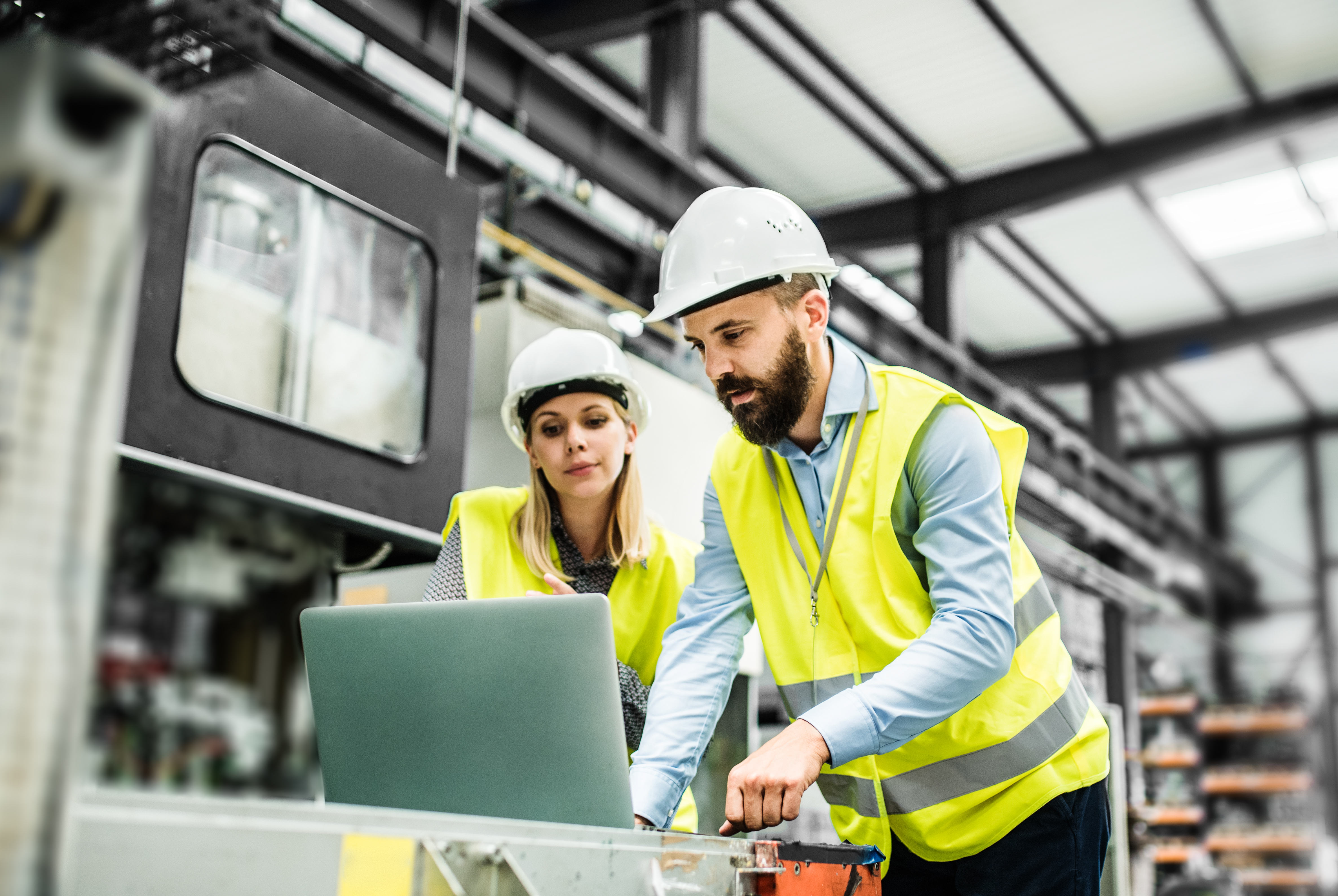Two construction workers in hard hats and yellow safety vests review plans on a laptop inside an industrial warehouse facility.