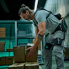 Worker in gray shirt and tool belt organizing cardboard boxes in a modern warehouse with teal shelving.