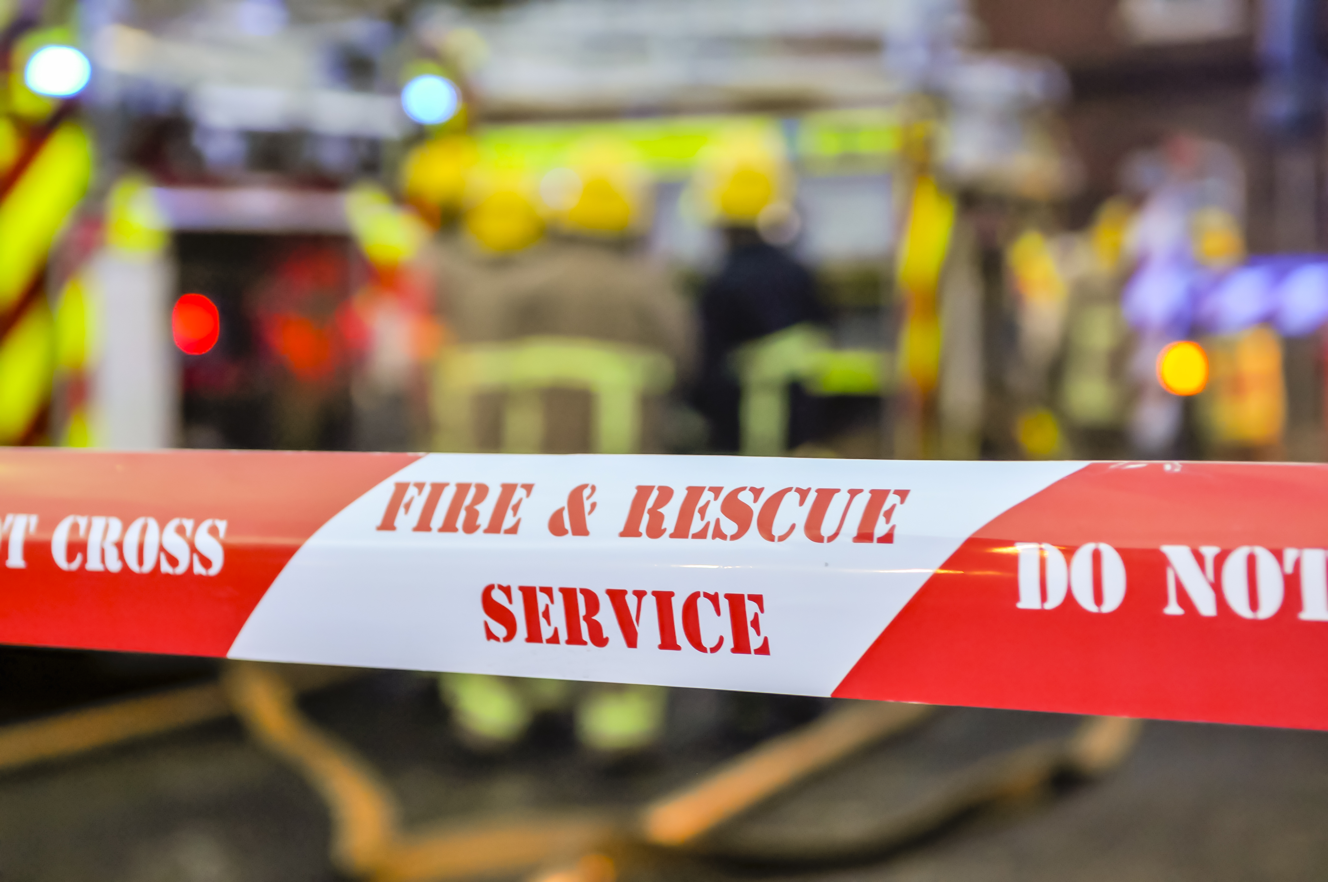 Red and white fire and rescue service barrier tape with 'Do Not Cross' warning at emergency scene with vehicles in background