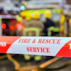 Red and white fire and rescue service barrier tape with 'Do Not Cross' warning at emergency scene with vehicles in background