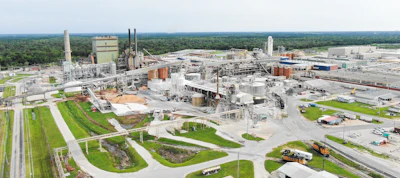 Aerial view of large industrial manufacturing facility with multiple buildings, smokestacks, and storage tanks surrounded by green landscape