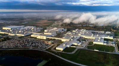 Aerial view of large industrial manufacturing complex with warehouse facilities, parking areas, and surrounding countryside landscape under cloudy sky