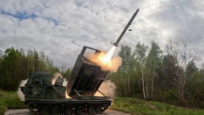 Military MLRS launcher firing a long-range missile in a forest clearing with smoke and flames visible
