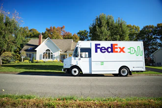 FedEx delivery truck parked on residential street in front of white house with autumn trees