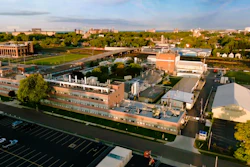 Aerial view of brick industrial complex with parking lot, green spaces, and surrounding cityscape