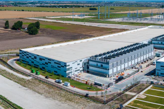 Aerial view of modern industrial warehouse complex with blue-accented buildings, loading docks, and surrounding agricultural landscape.
