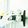 Gloved hand holding a brass gas regulator valve with pressure gauges and black control knob in laboratory setting