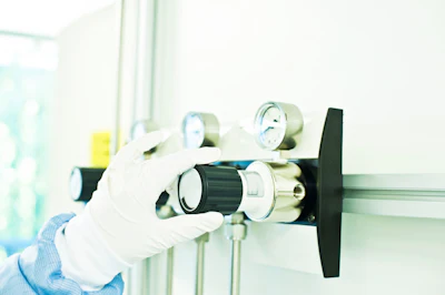 Gloved hand holding a brass gas regulator valve with pressure gauges and black control knob in laboratory setting