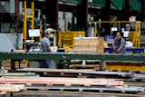 Workers processing lumber at an industrial woodworking facility with yellow machinery and green structural beams.