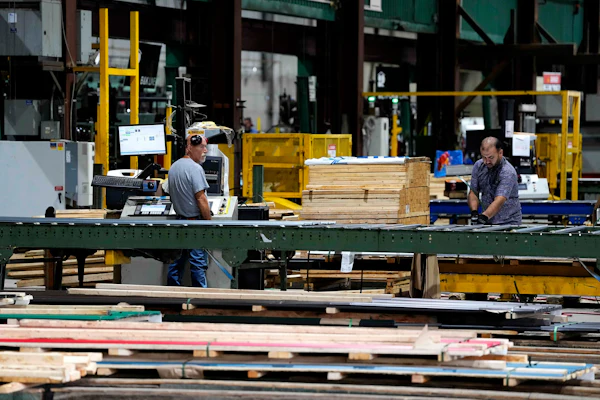 Workers processing lumber at an industrial woodworking facility with yellow machinery and green structural beams.