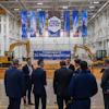 Corporate event in industrial warehouse with blue banner reading 'Future Home of 24 Hadrian Machines' and yellow excavators