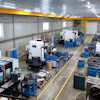 Industrial manufacturing facility with CNC machines, blue workbenches, and overhead yellow crane system in warehouse space