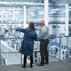 Two professionals observe a large industrial manufacturing facility from an elevated platform with expansive windows.