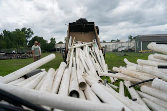 White PVC pipes being unloaded from a dump truck at an industrial yard with green buildings in background