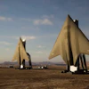 Three large metallic sail-shaped sculptures stand in a desert landscape under a blue sky with clouds