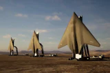 Three large metallic sail-shaped sculptures stand in a desert landscape under a blue sky with clouds