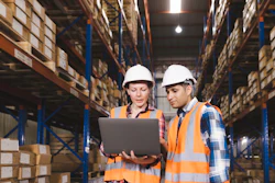 Two warehouse workers in safety gear reviewing inventory data on laptop in storage facility
