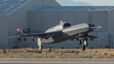 Silver and black experimental aircraft landing near industrial hangar with mountains in background